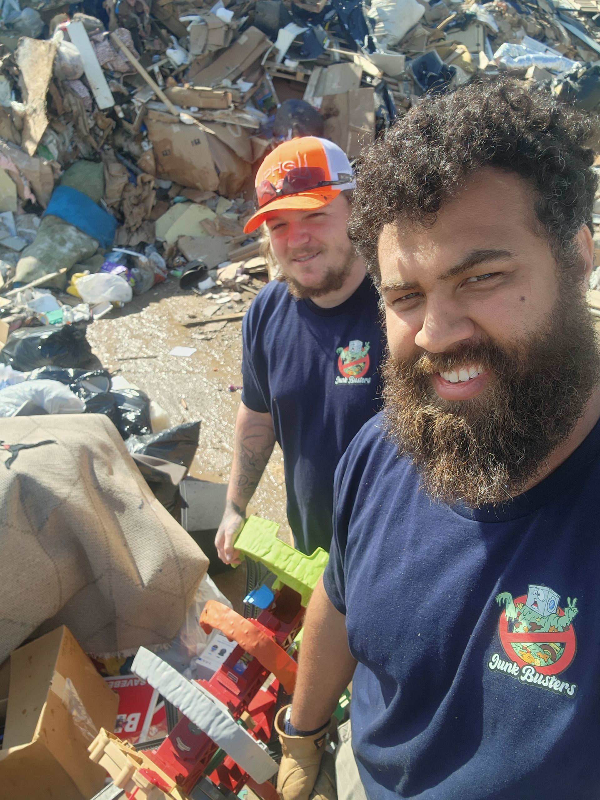 Two men smile, wearing matching blue shirts, amidst a pile of debris. One man has a beard.