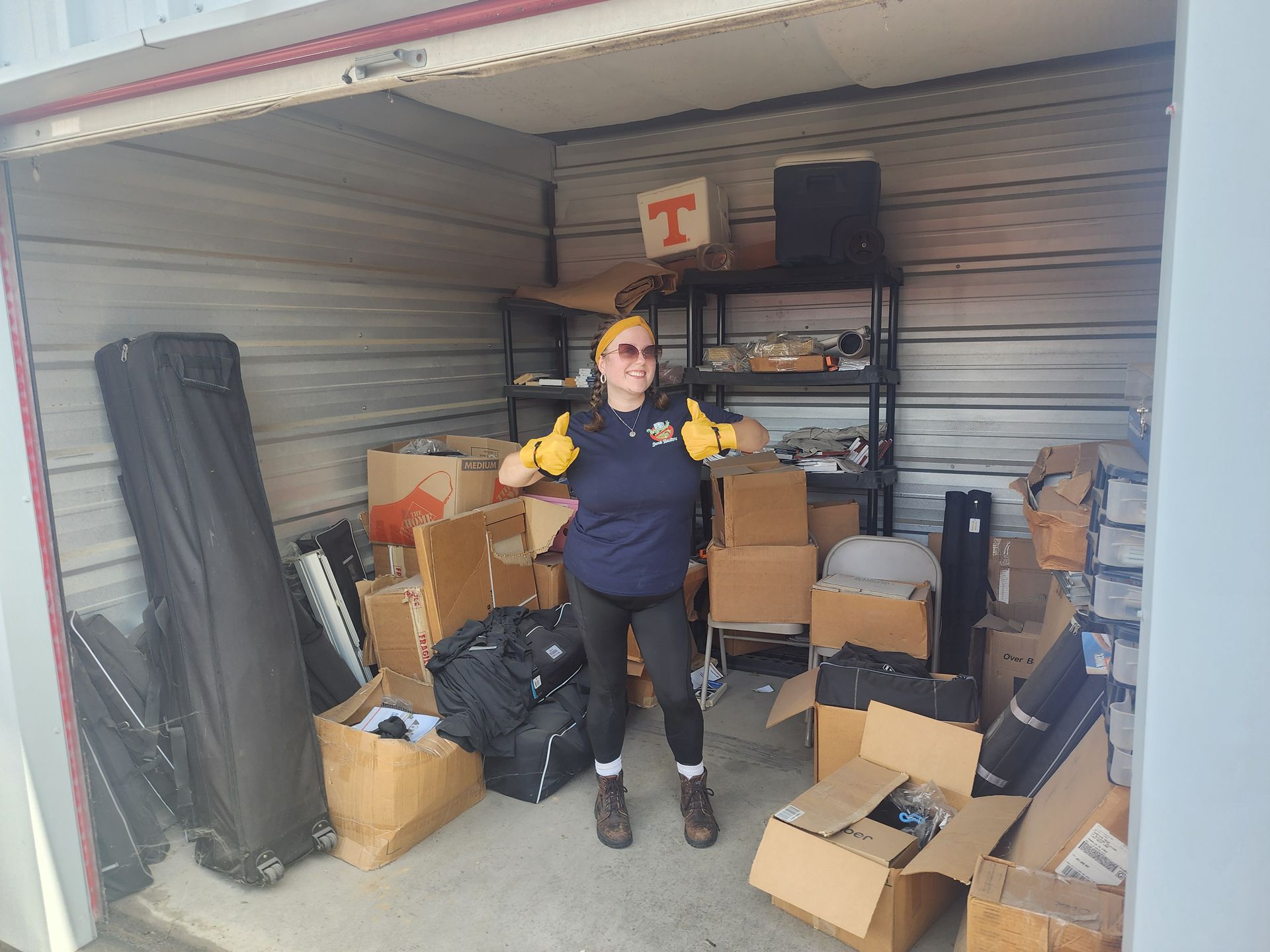 Woman in storage unit surrounded by boxes, thumbs up.