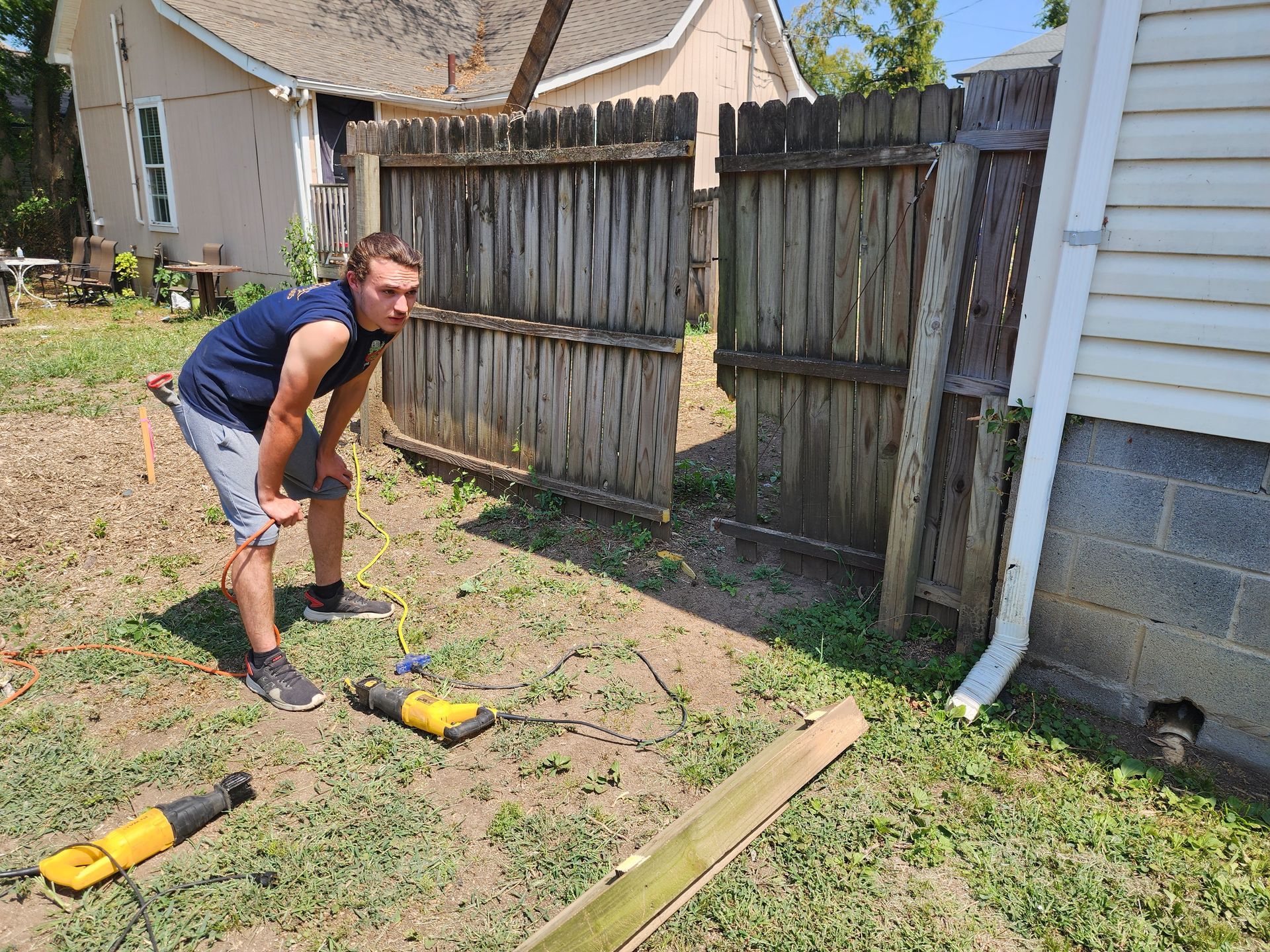 Man leaning over, working on a wooden fence outdoors, two yellow tools on the ground.