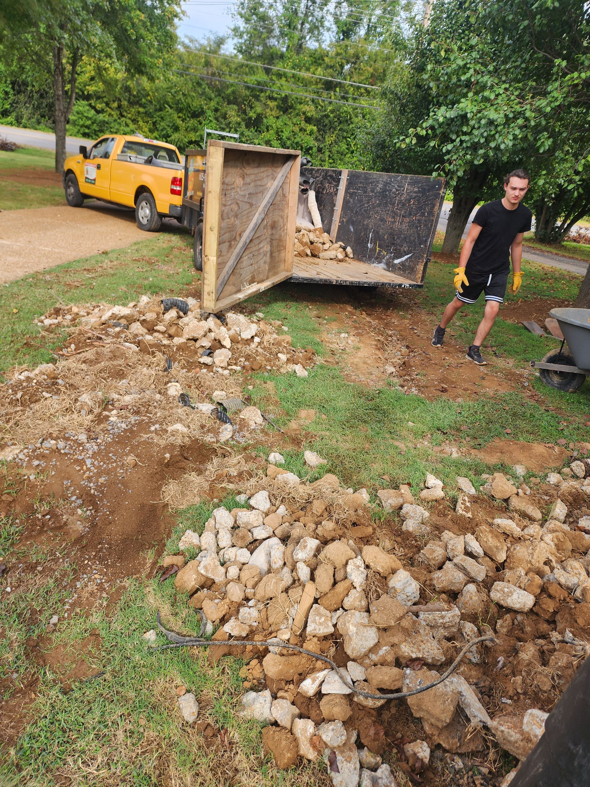 Man unloading rocks from a trailer. Yellow truck and trailer on grass, rocks on the ground.