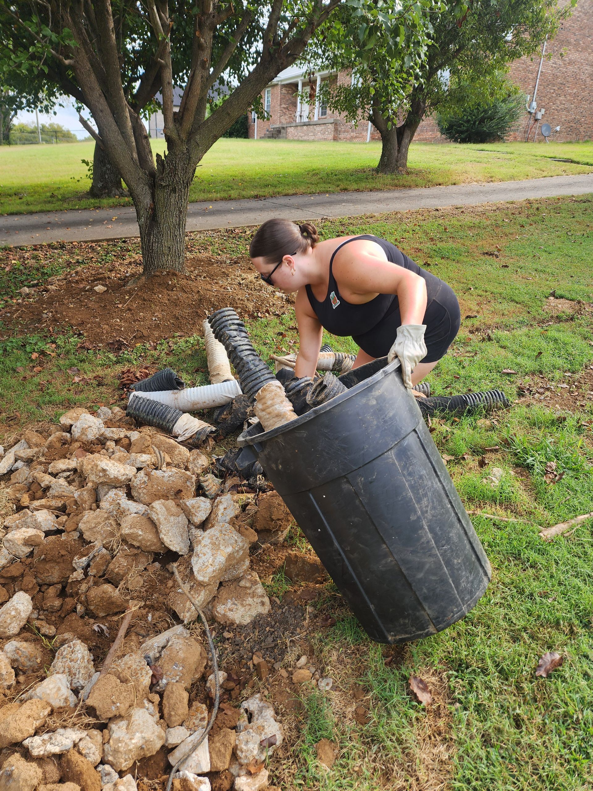 Woman in black tank top and shorts emptying a black bin of rocks and debris outdoors.