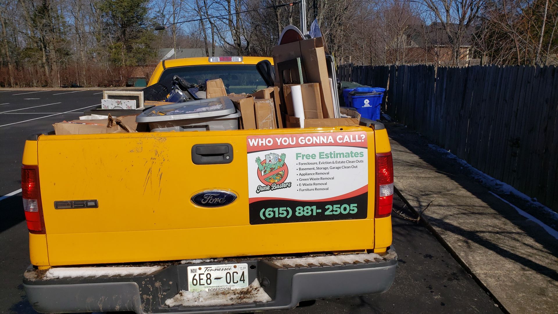 Yellow truck bed loaded with boxes and items, parked next to a fence. License plate is visible.