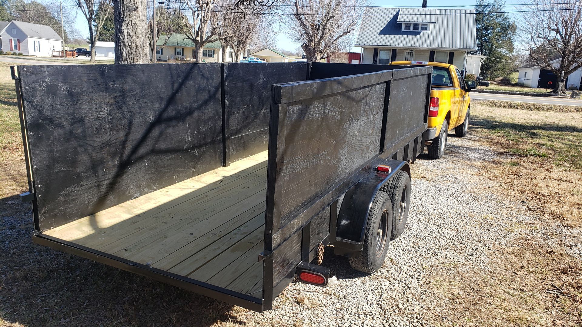 Black utility trailer hitched to a yellow truck, parked outside on gravel, in front of houses.