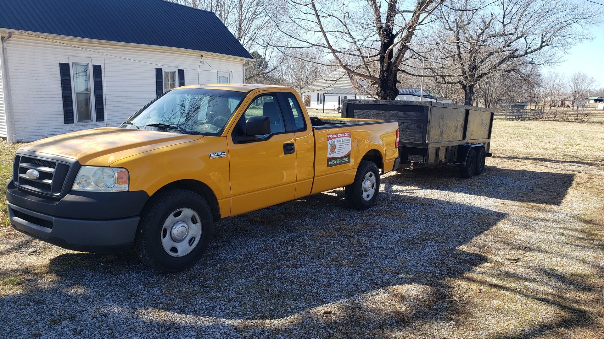 Yellow pickup truck towing a trailer on gravel; near a house and tree.