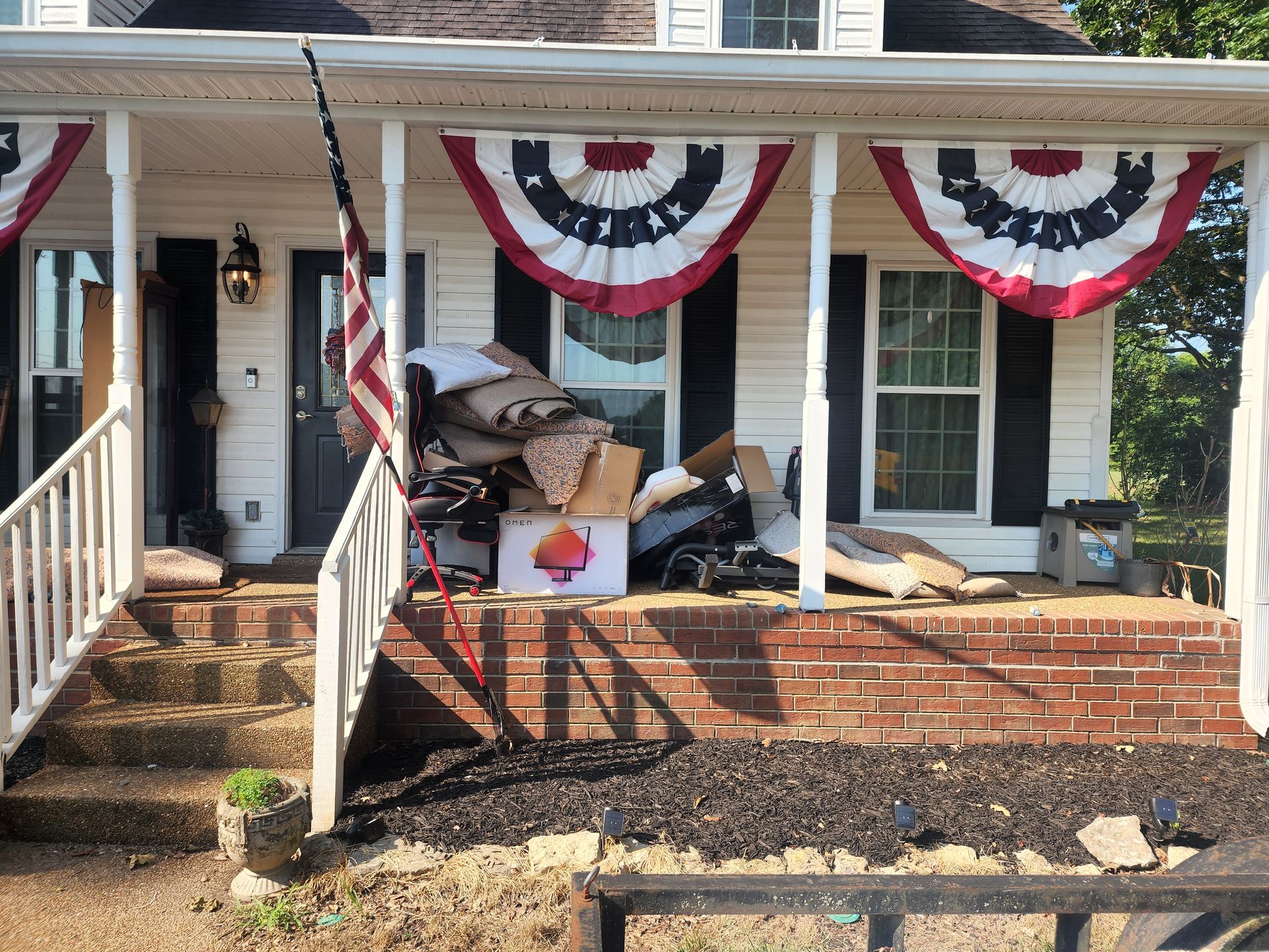 Front porch of a house with moving supplies: boxes, furniture, and American flag. Red, white, and blue bunting.