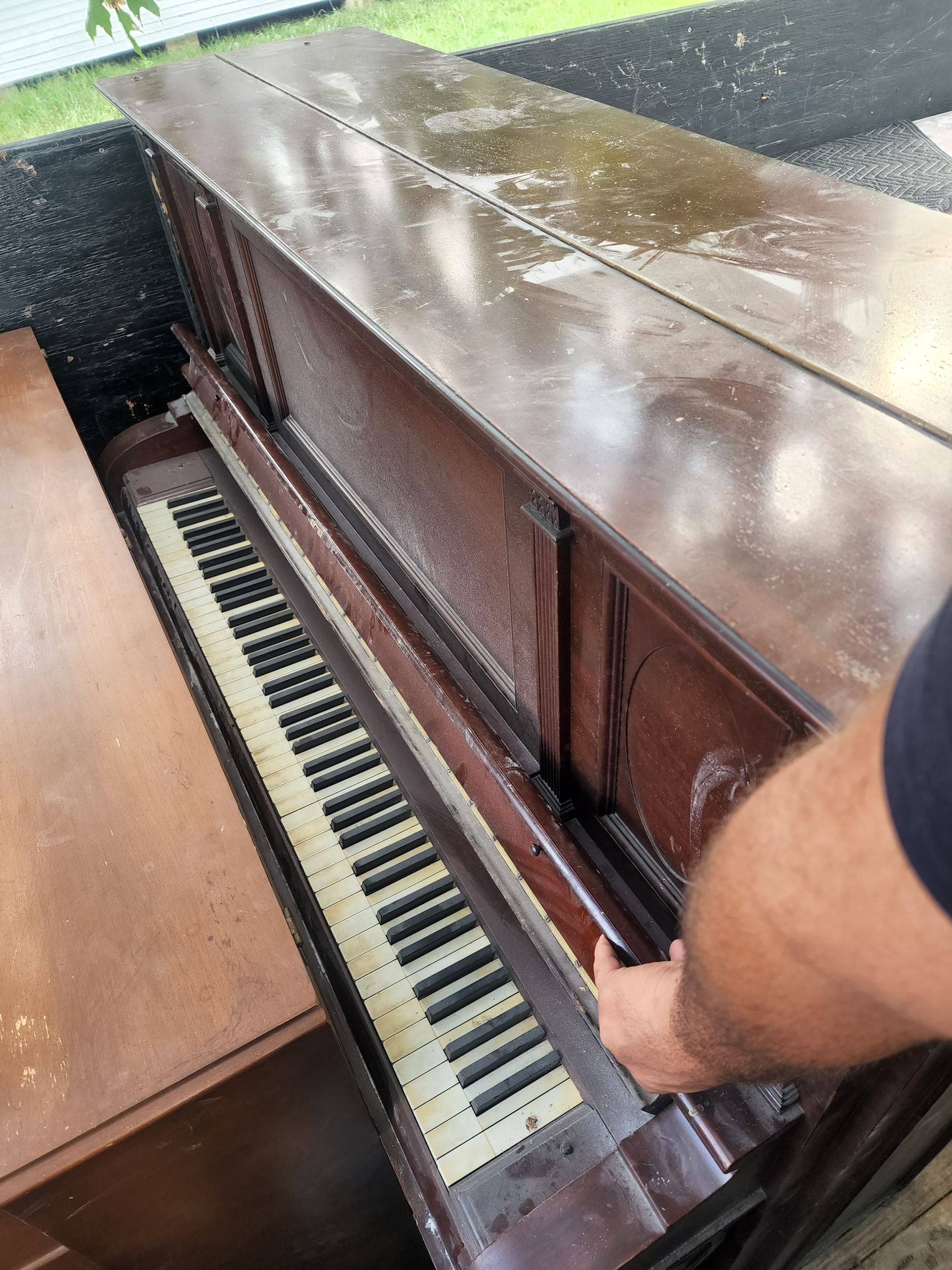 Man holding a damaged piano; brown wood, keys visible, outdoors.
