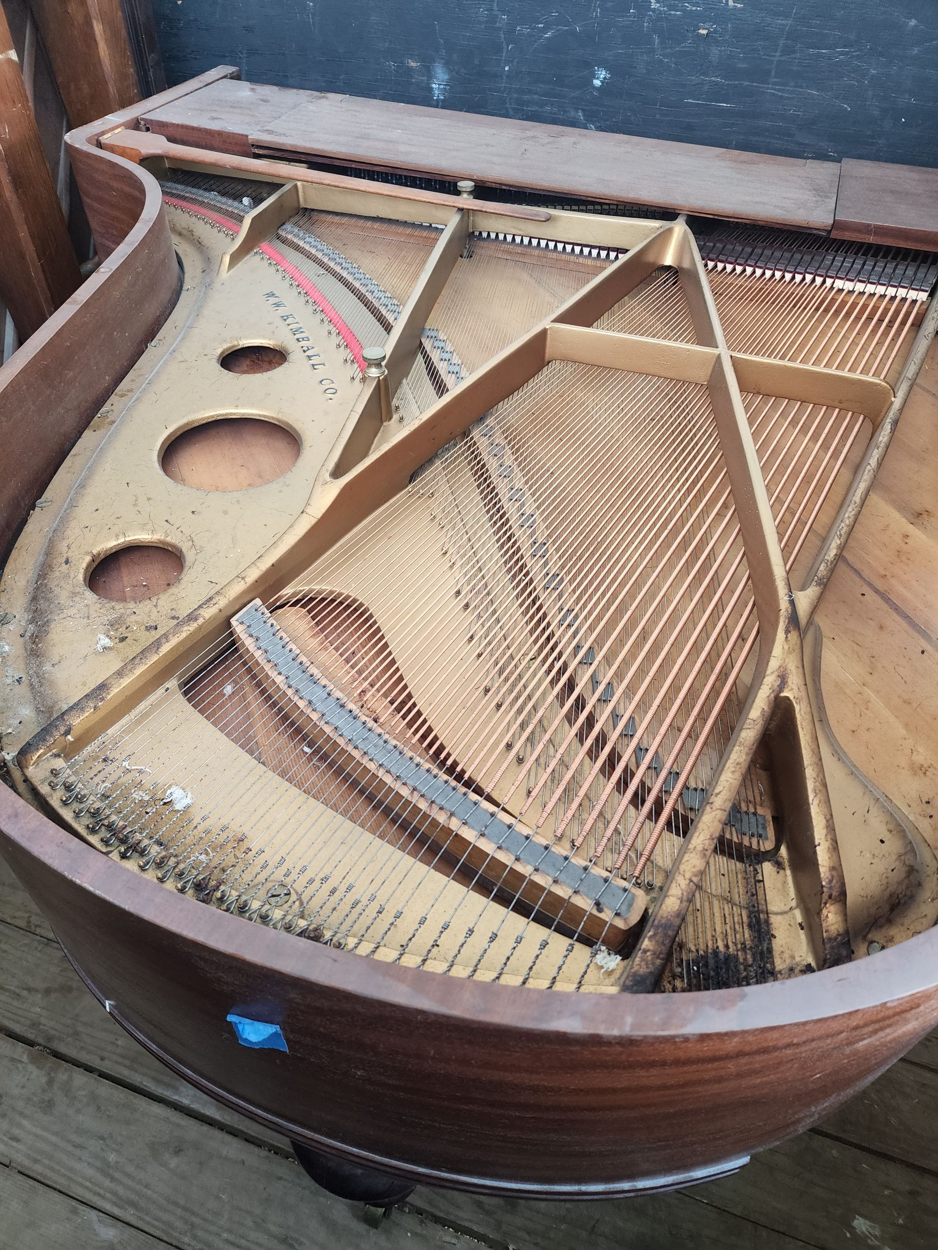 Open grand piano, strings exposed, wooden frame visible. Brown and tan tones, outdoors.