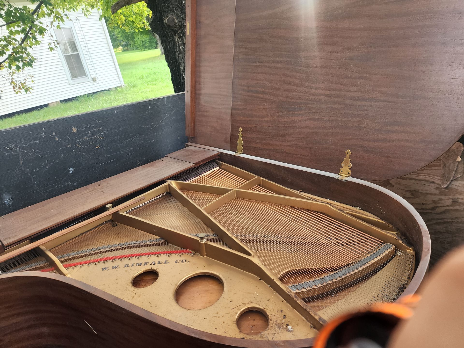 Open grand piano, strings and soundboard visible, outdoors with sunlight and a house in the background.