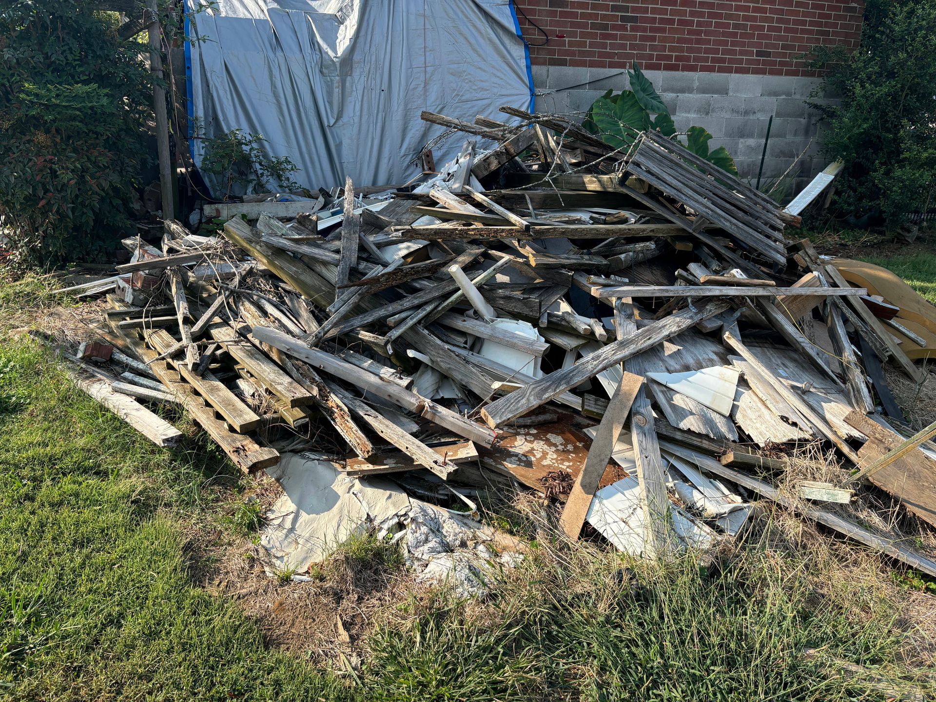 Pile of weathered wood debris on grass near a brick building, covered by a blue tarp.