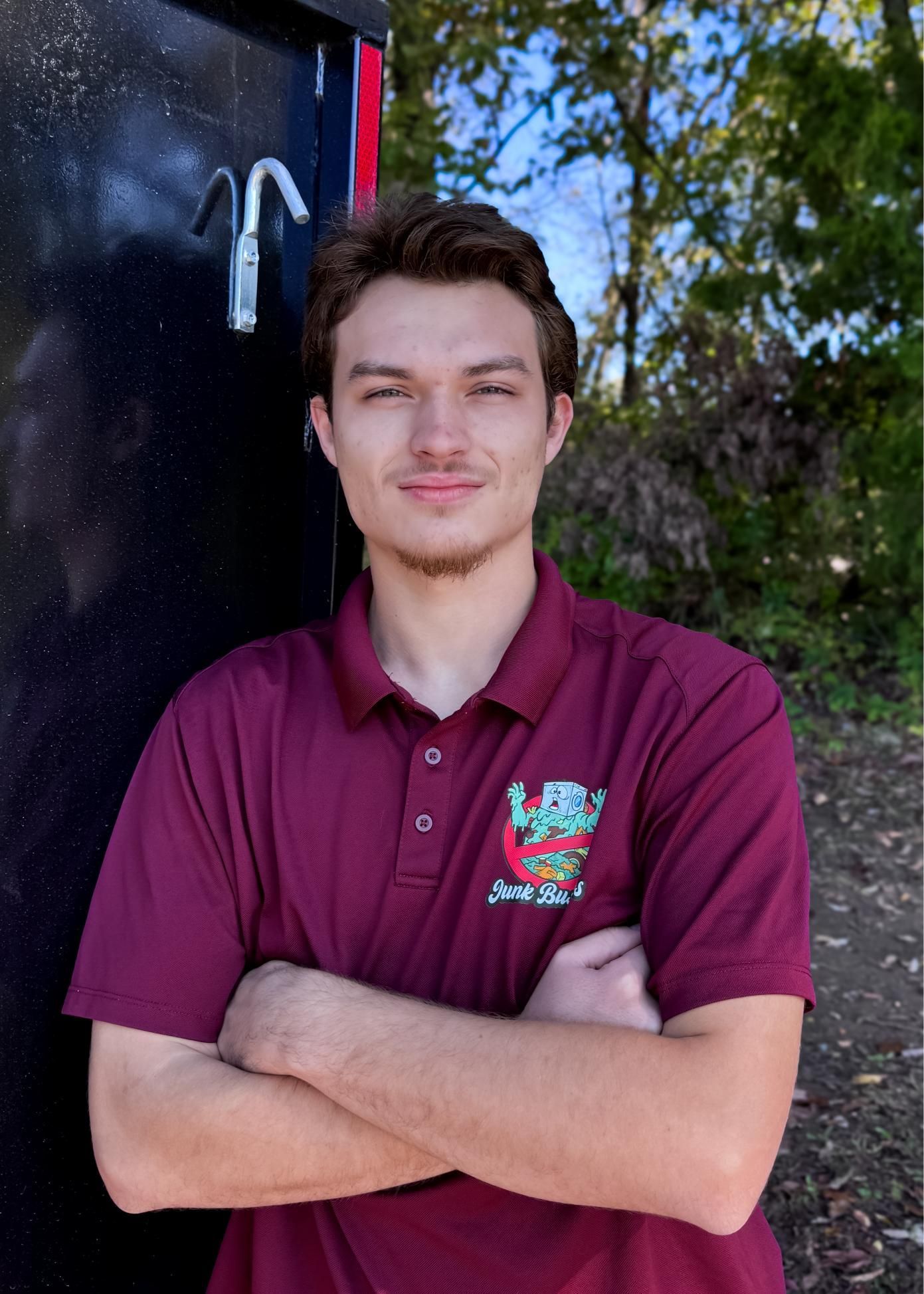 Man with arms crossed, wearing maroon polo shirt with logo, standing outdoors.