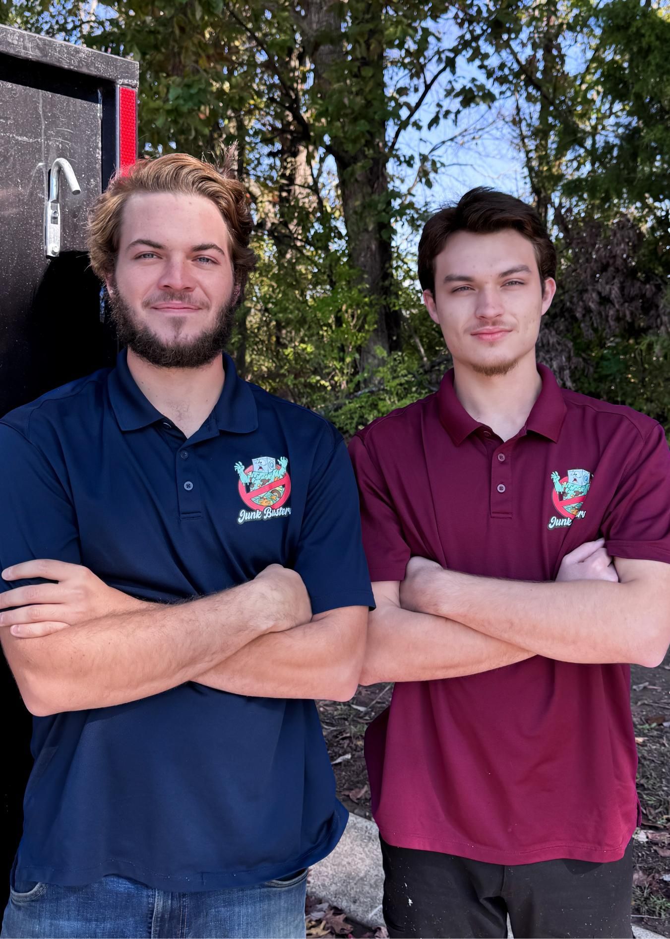 Two men in matching polo shirts stand outdoors with arms crossed.