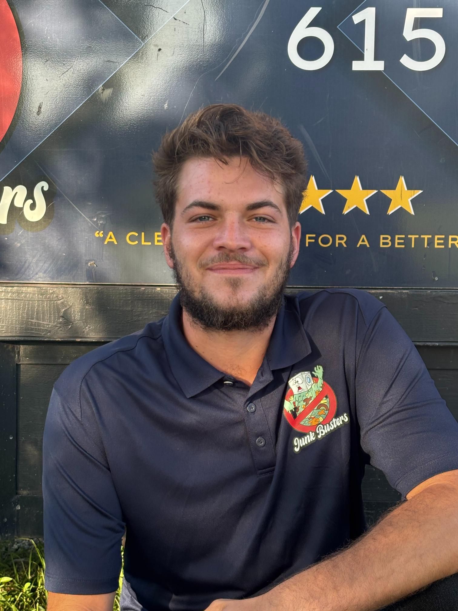 Man in a dark blue polo shirt smiles, sits outdoors; background: 