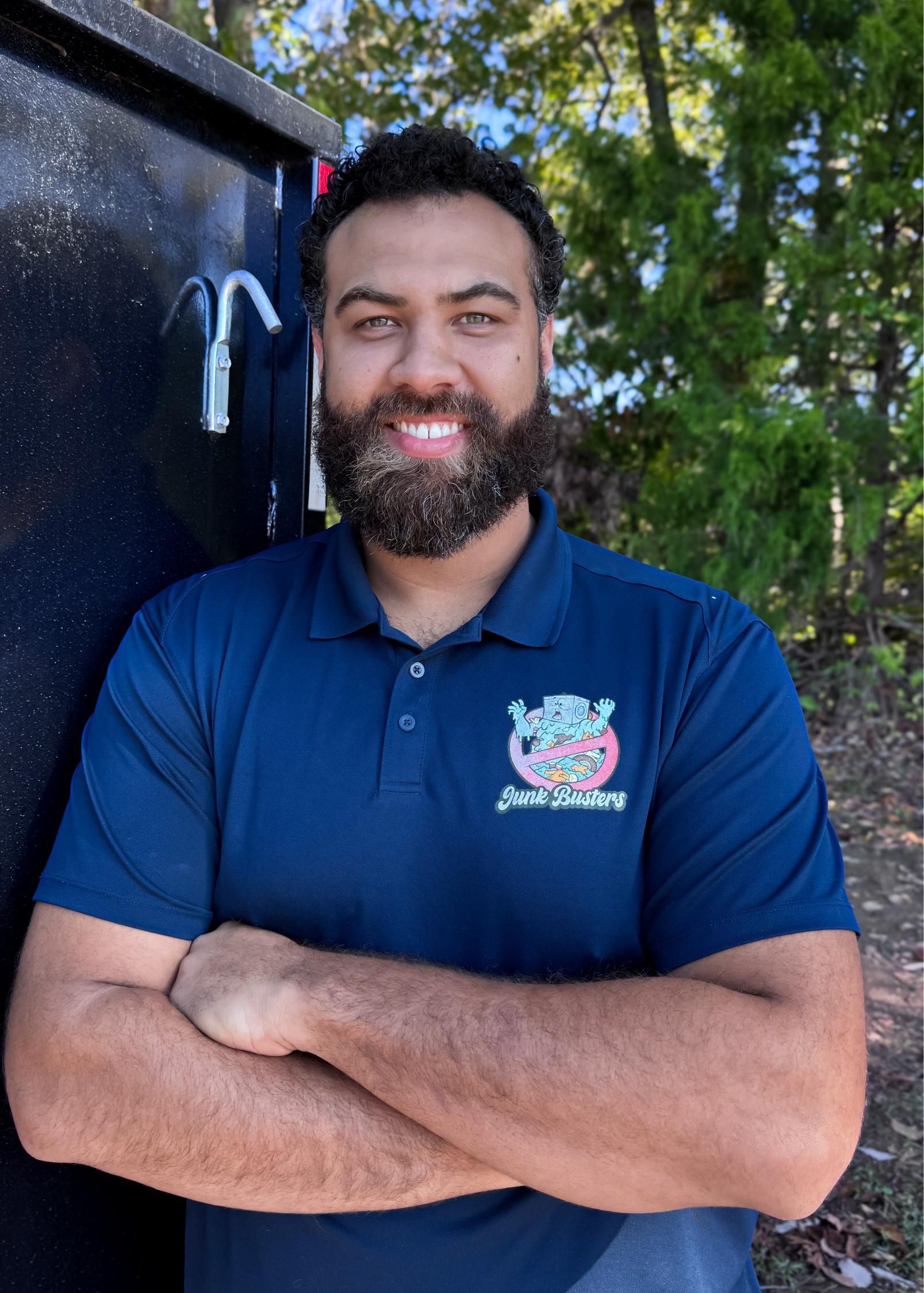 Man with beard, arms crossed, smiling. Wearing a blue polo shirt, standing near a dark container, outdoors.