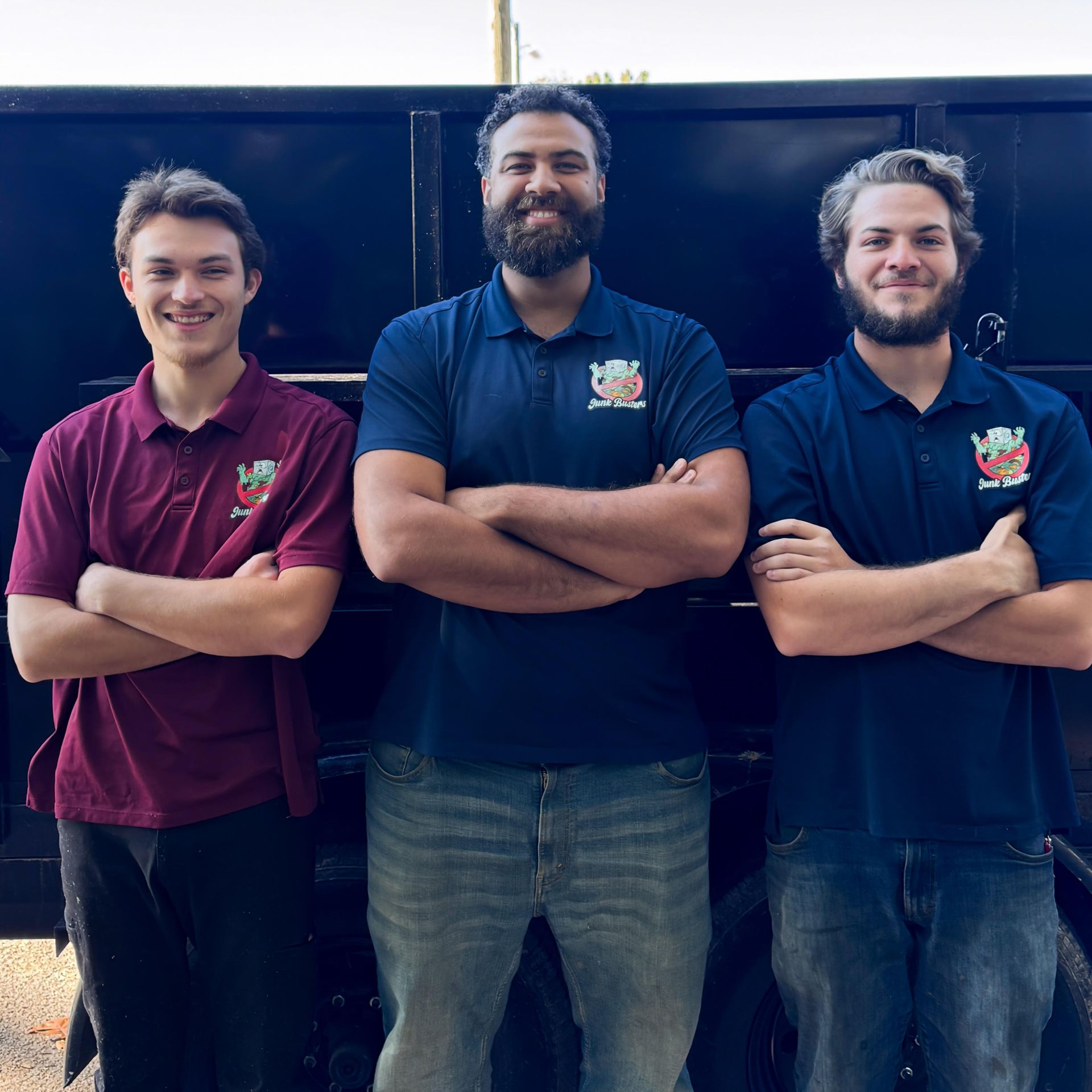 Three men with arms crossed, in front of a dark truck. They wear polo shirts, jeans, smiling.
