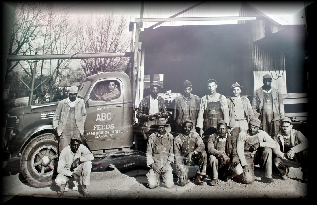 A group of fourteen workers in overalls pose in front of a parked truck labeled