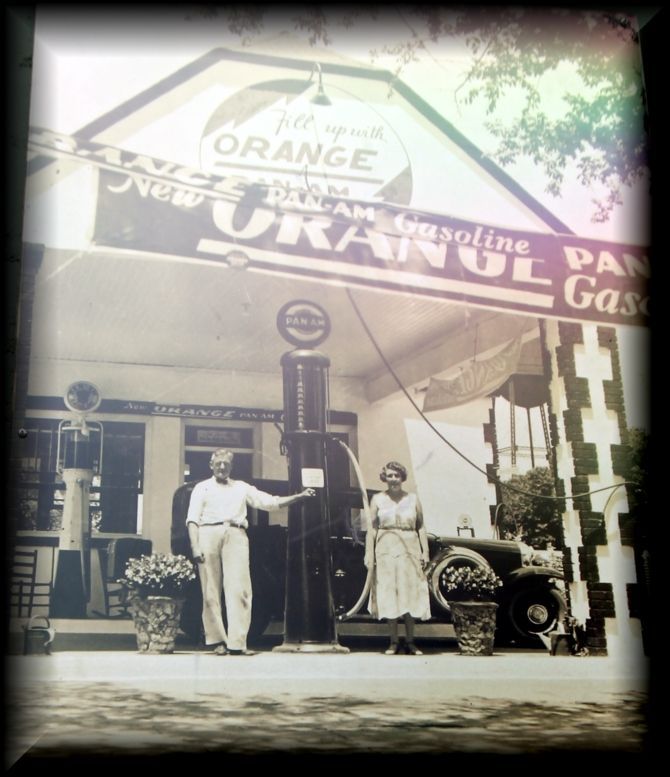 A man and woman stand at a Pan-Am service station in front of a vintage gas pump and parked car.