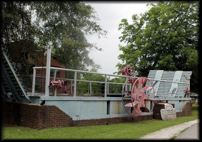A large, weathered light-blue mechanical lock gate stands over a brick foundation in a park with green grass and trees.