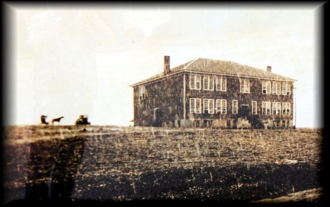 A two-story, rectangular school building stands on a barren, plowed field with a horse and carriage nearby.