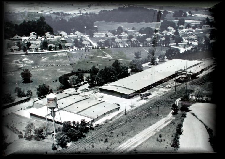 An aerial, black-and-white view of an industrial factory complex with a water tower, adjacent to a small residential town.