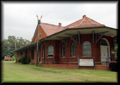 A red brick train depot with a terracotta tile roof and wide eaves, situated in a green, grassy area under a cloudy sky.