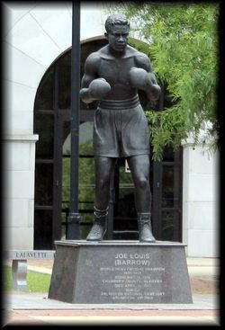 A bronze statue of a boxer in a fighting stance on a stone pedestal, located outdoors in front of a building.