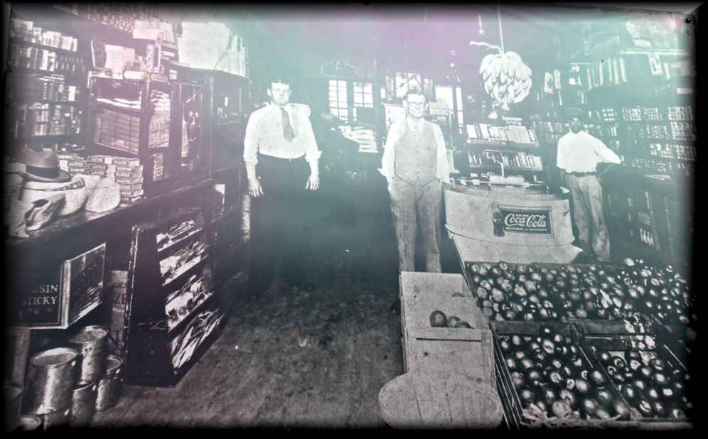 A vintage black-and-white photo of three people standing inside a general store filled with merchandise and produce.