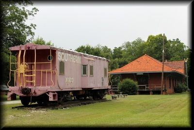 A pink Southern Railway caboose sits on train tracks next to a historic brick train depot with a red tiled roof.