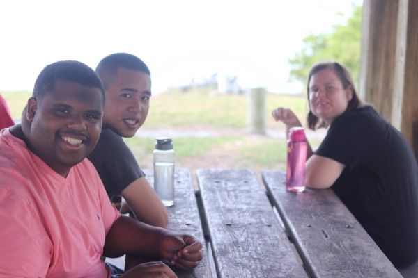 Three people seated at a picnic table outdoors, smiling toward the camera with water bottles on the table.