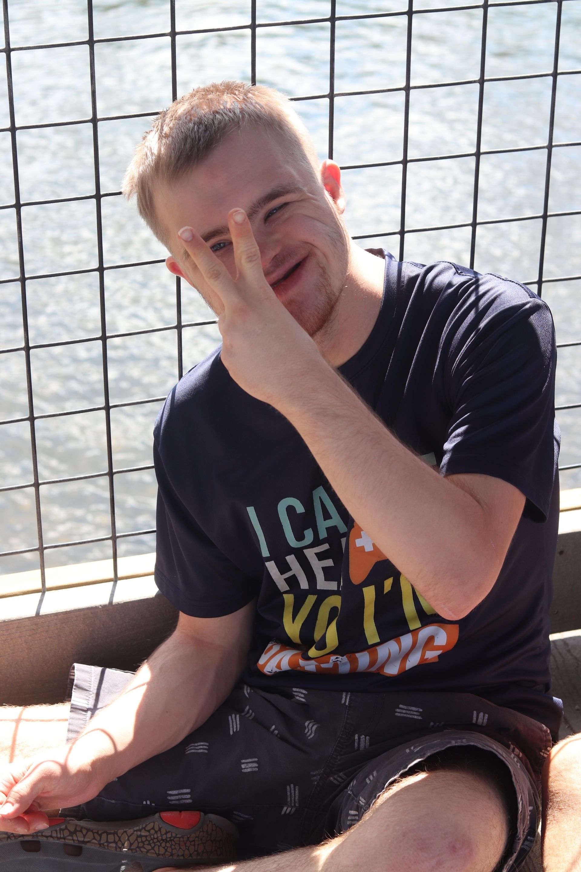 Person sitting by a wire fence, smiling and covering their face with one hand.