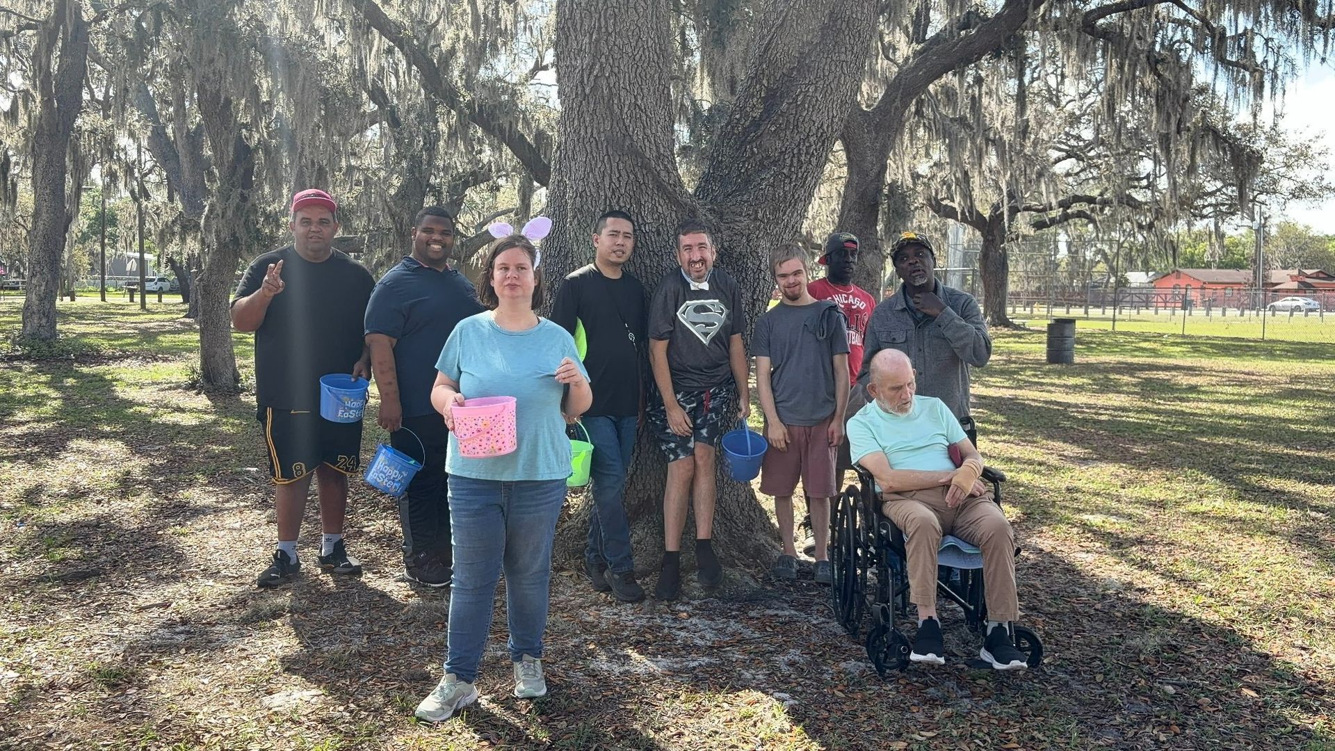 Group of people standing and sitting outdoors in a wooded park, holding colorful papers.