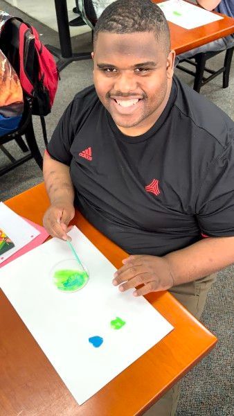 Person smiling at a classroom desk, using green paint on white paper with colorful shapes.