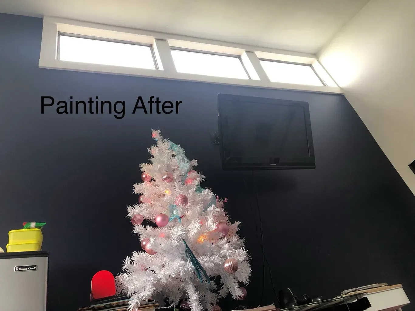 A dark blue painted wall with a white Christmas tree and a mounted TV. White windows are above the TV.