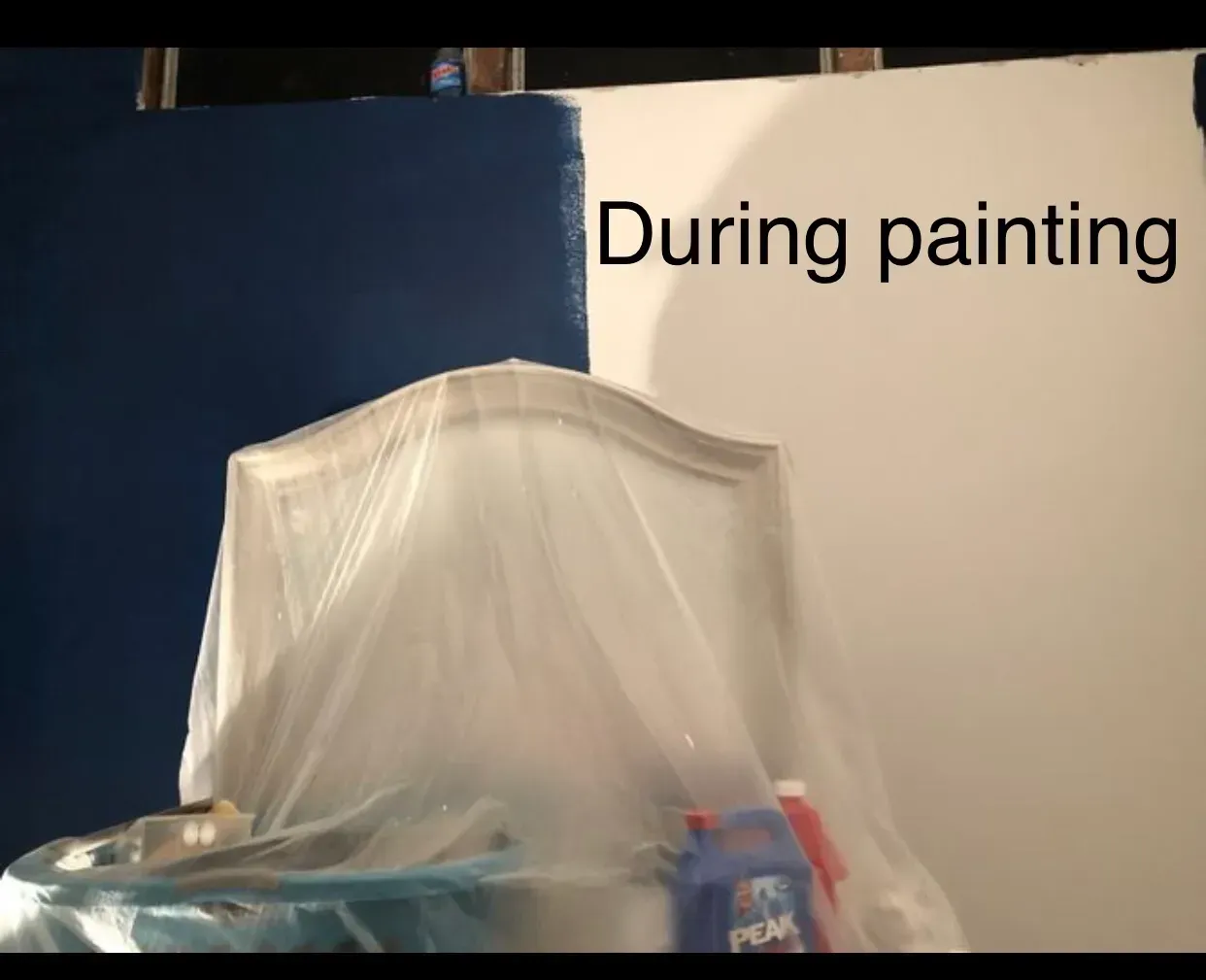 Wall partially painted blue with a headboard draped in plastic and painting supplies.