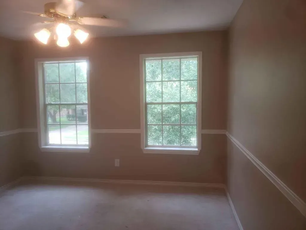 Empty beige-walled room with two windows and a ceiling fan. White trim along walls and windows.