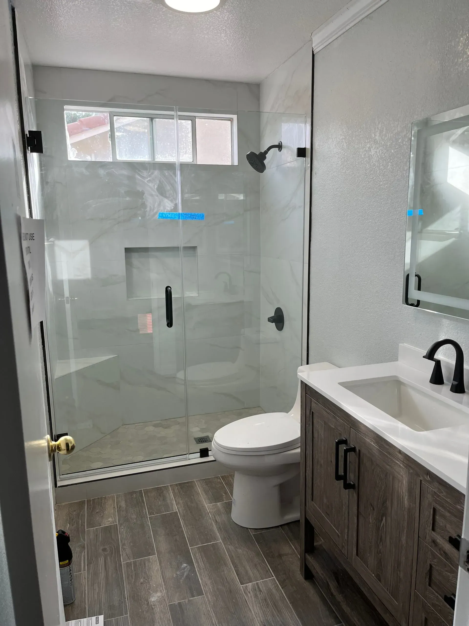 Bathroom with glass shower, wooden vanity, gray and white tile, and wood-look flooring.