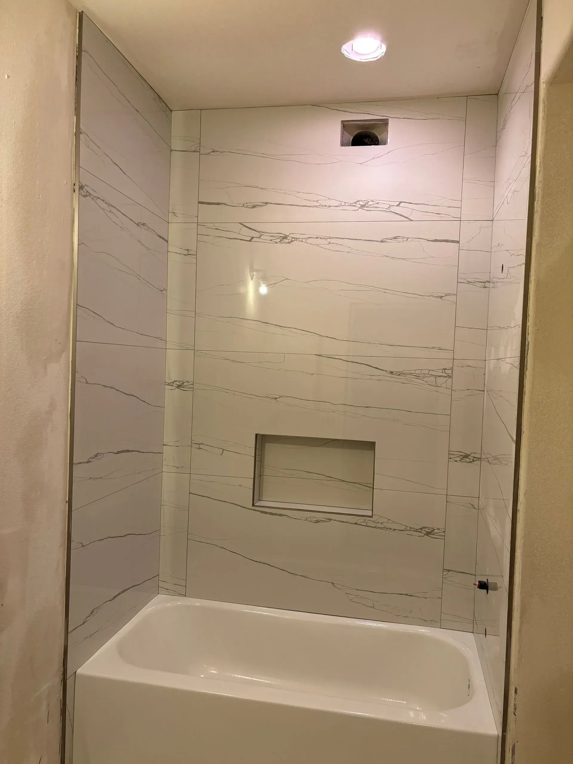 White and gray-veined shower surround with built-in shelf over white bathtub. Ceiling light and air vent are visible.
