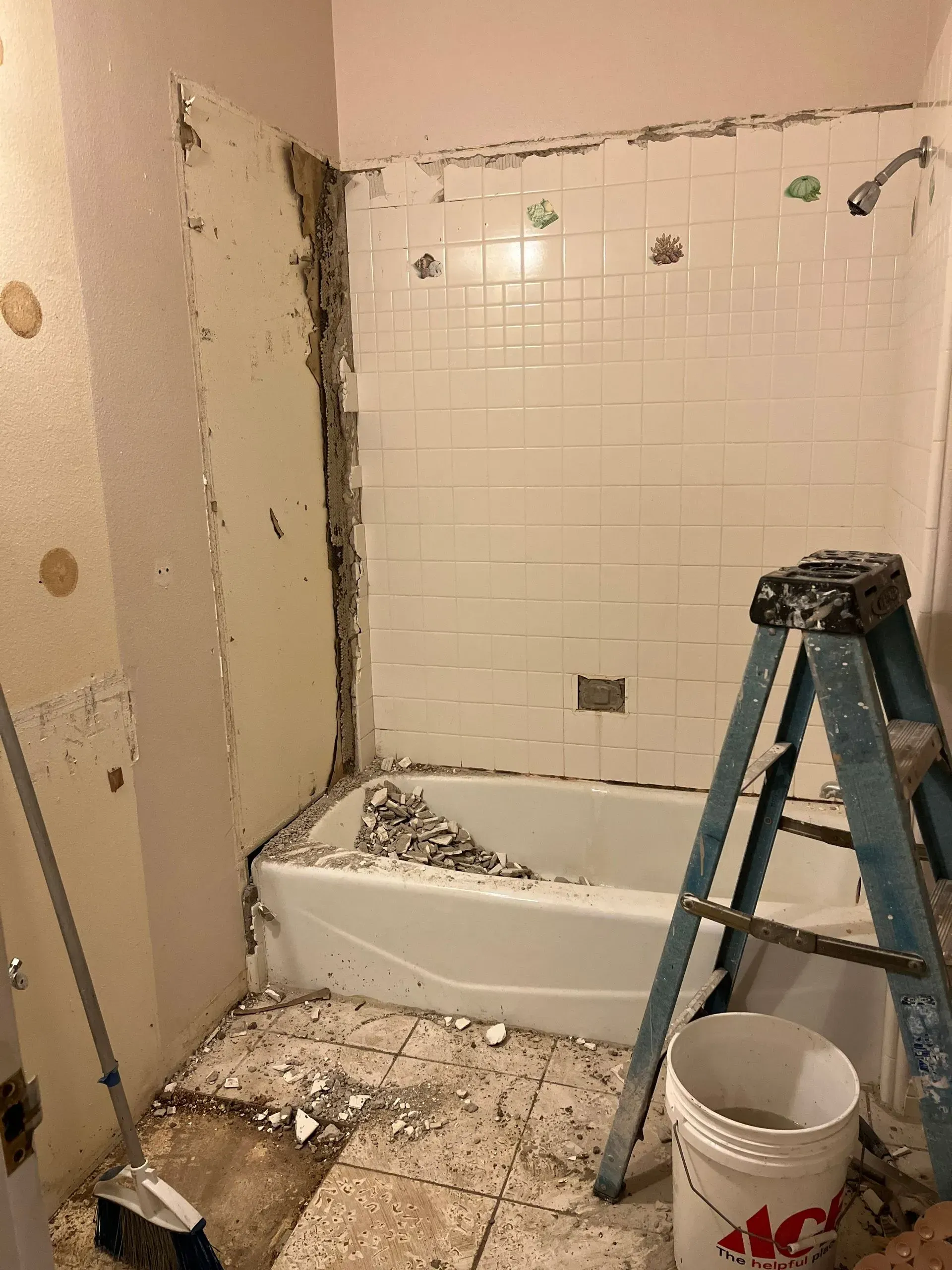 Bathroom under renovation, with tub, tile partially removed, drywall exposed, and ladder. Debris on the floor.