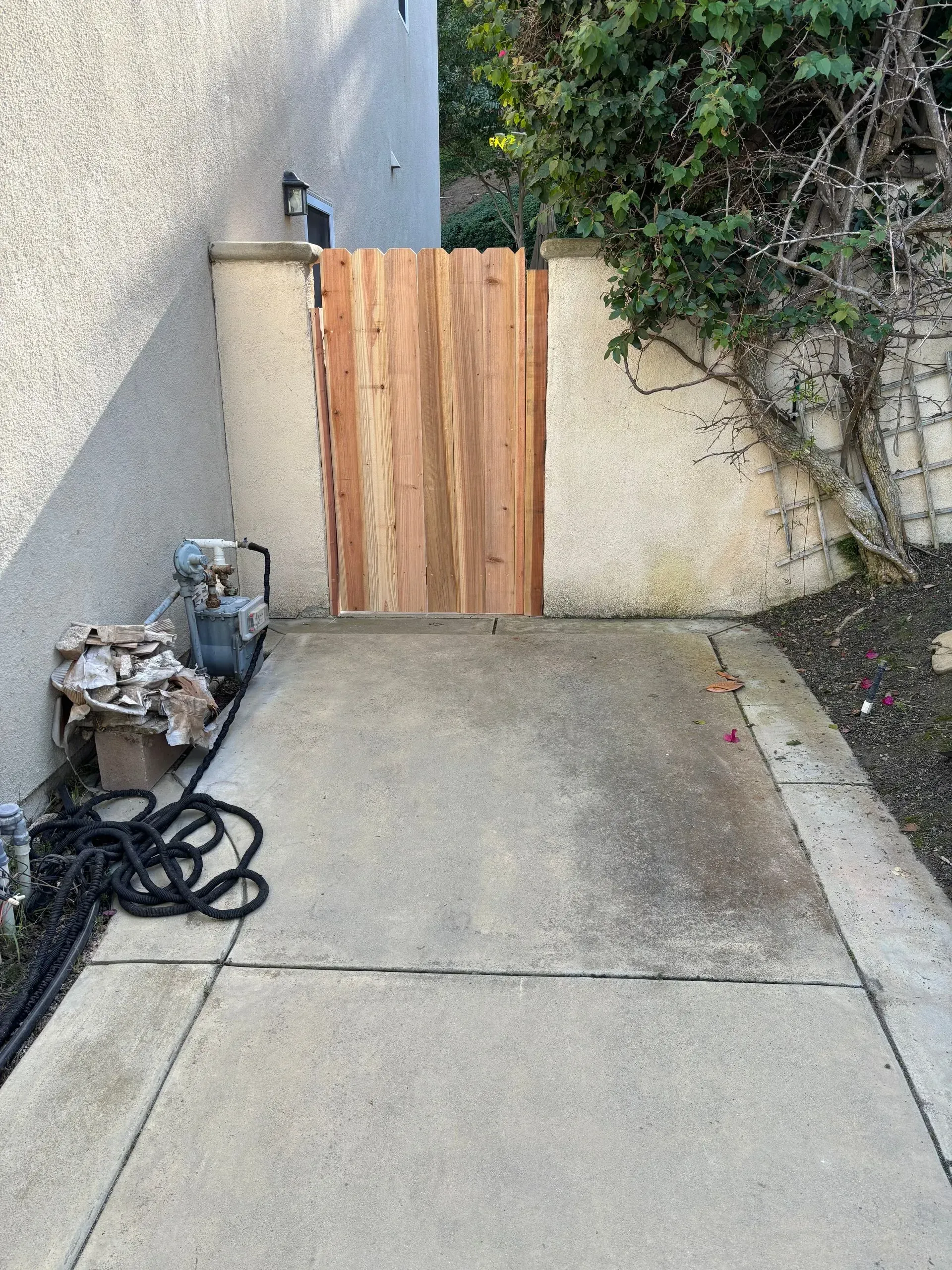 Concrete pathway leading to a wooden gate between textured walls; coiled black hose and utilities on left.