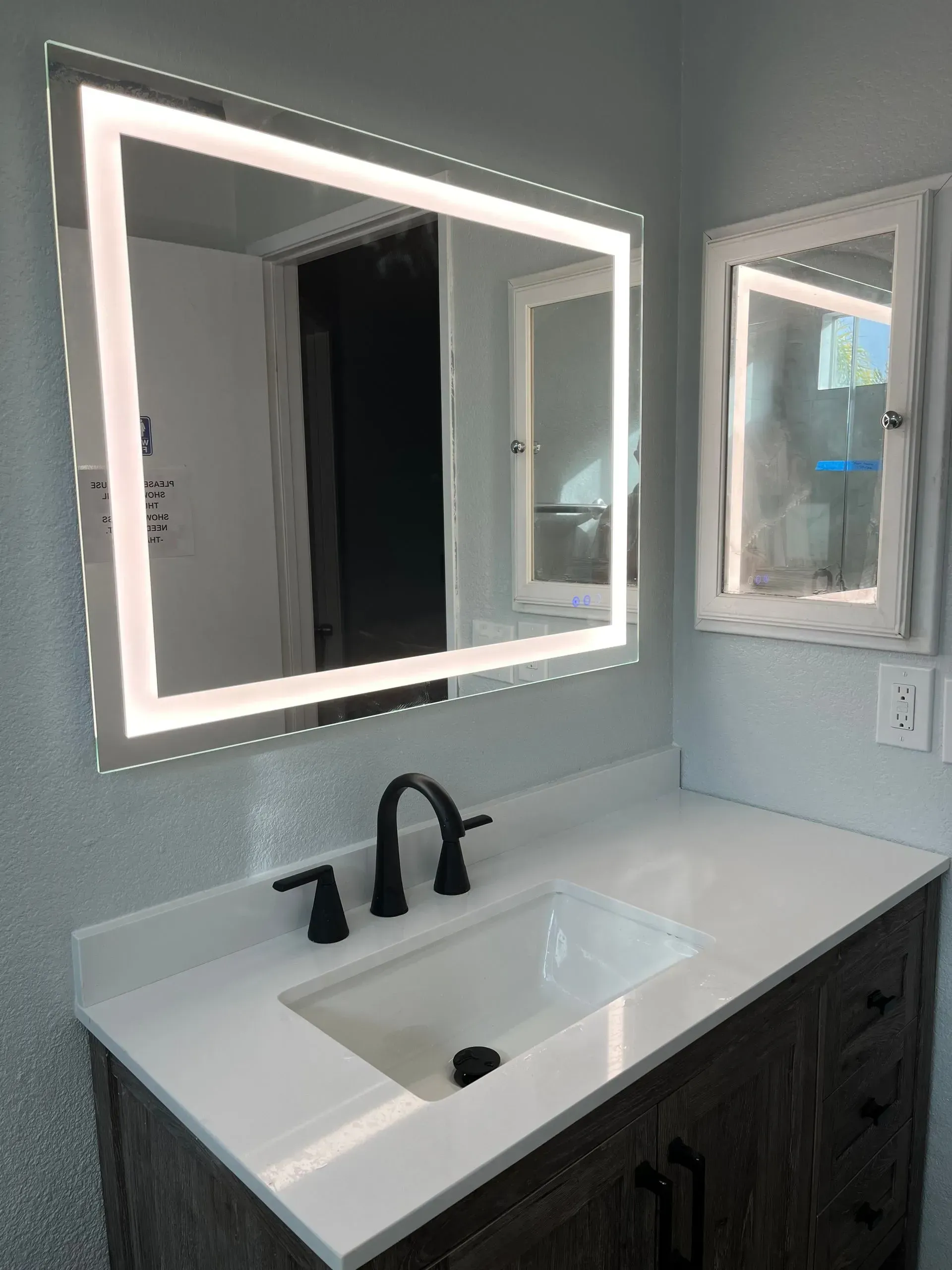 Bathroom with illuminated mirror, white countertop, dark faucet, and vanity.