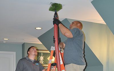 Two men are cleaning a ceiling with a broom while standing on a ladder.