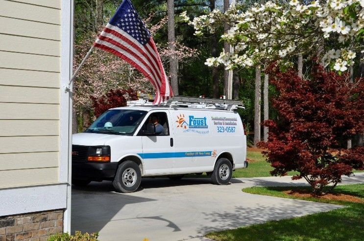 A white HVAC service van and ladder parked in a driveway near trees and an American flag