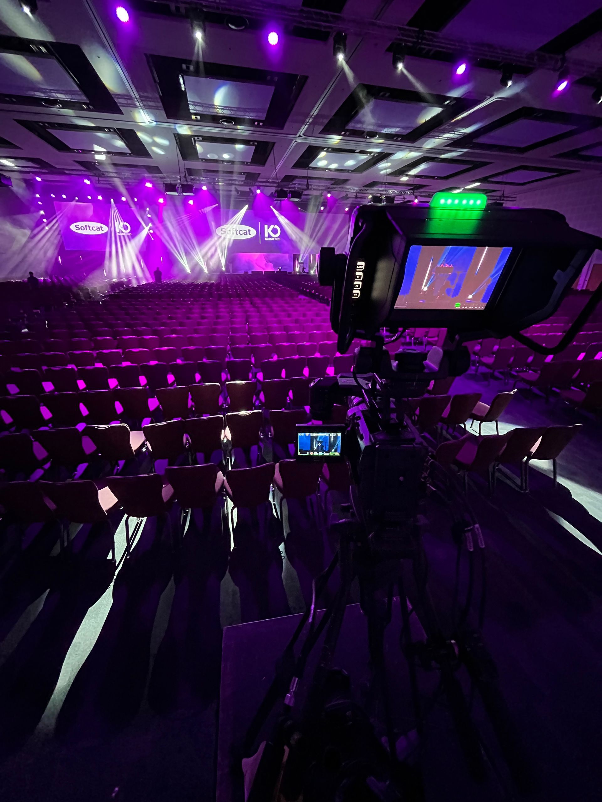 A camera on a tripod filming a brightly lit stage in a large venue, ready for a conference.