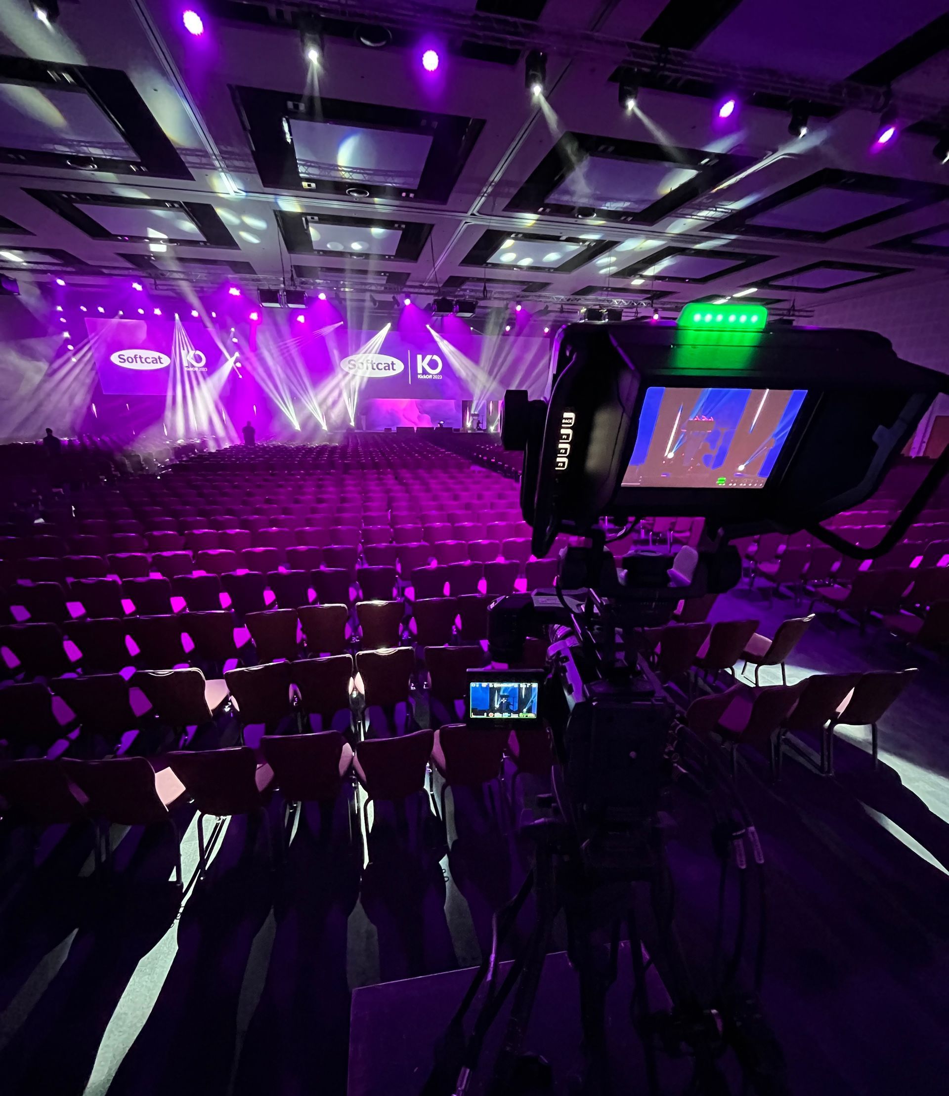Camera filming an event in a large venue with purple stage lights and seated audience.