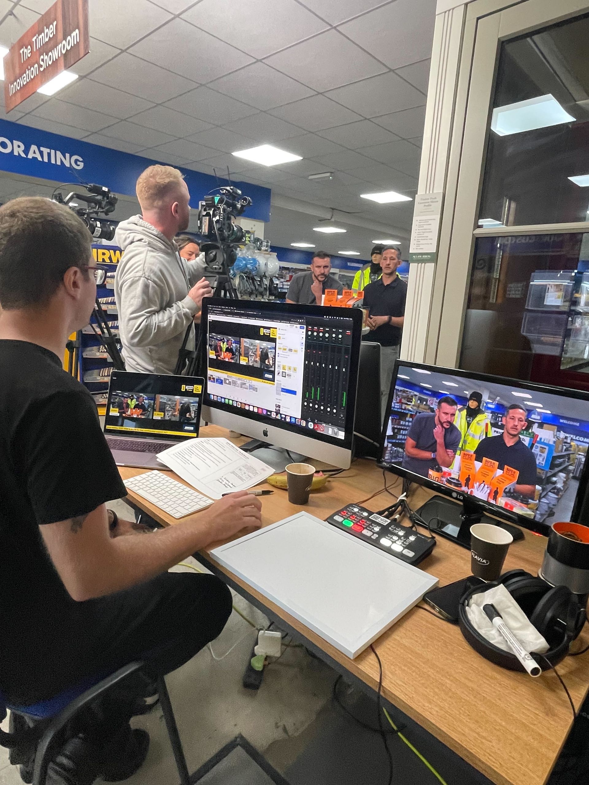 A live broadcast setup in a store. Crew members operate cameras and monitors. One man speaks into a microphone.
