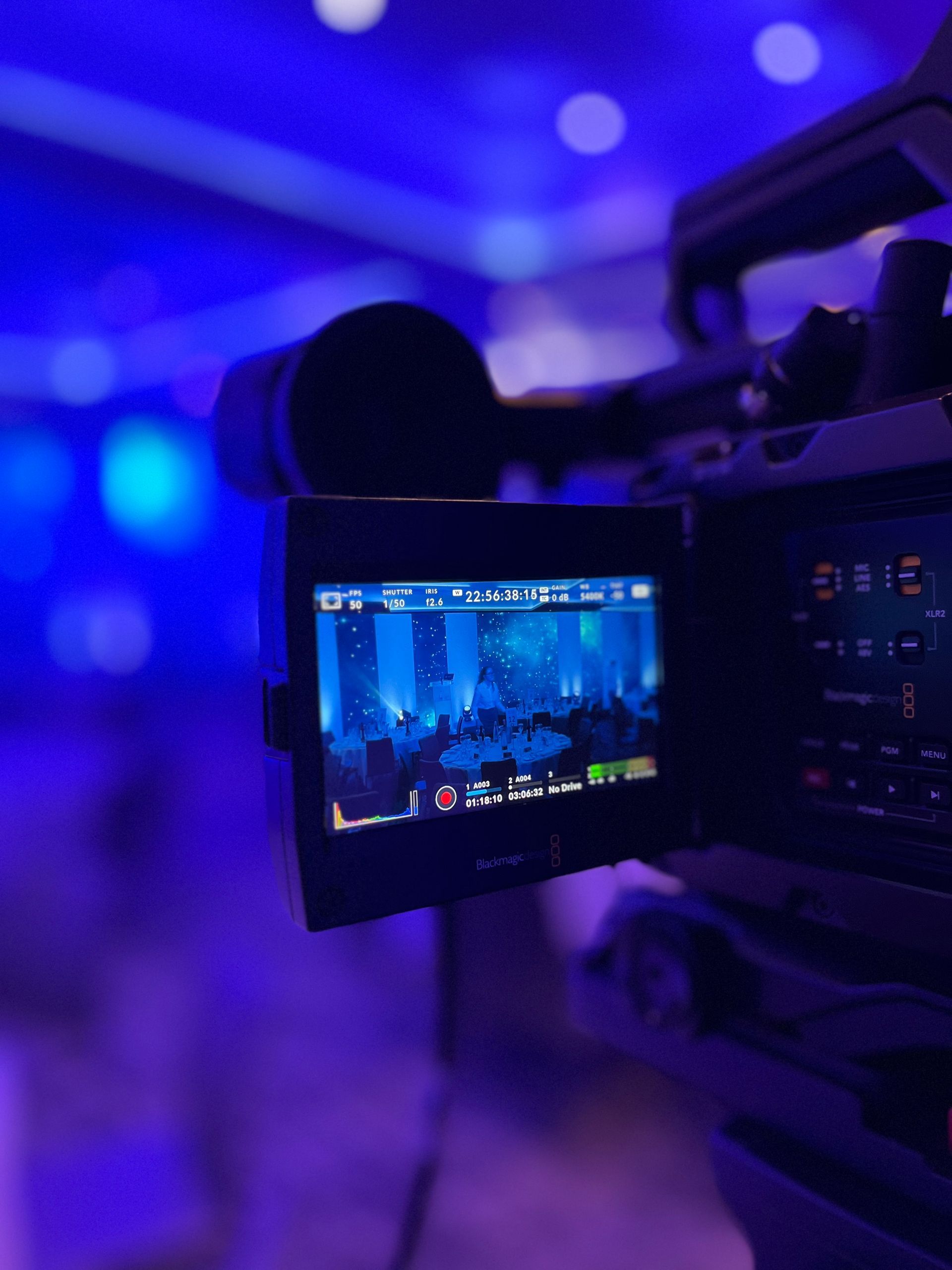 Close-up of a video camera recording an event in a room with blue lighting. The screen shows a live view of the setting.