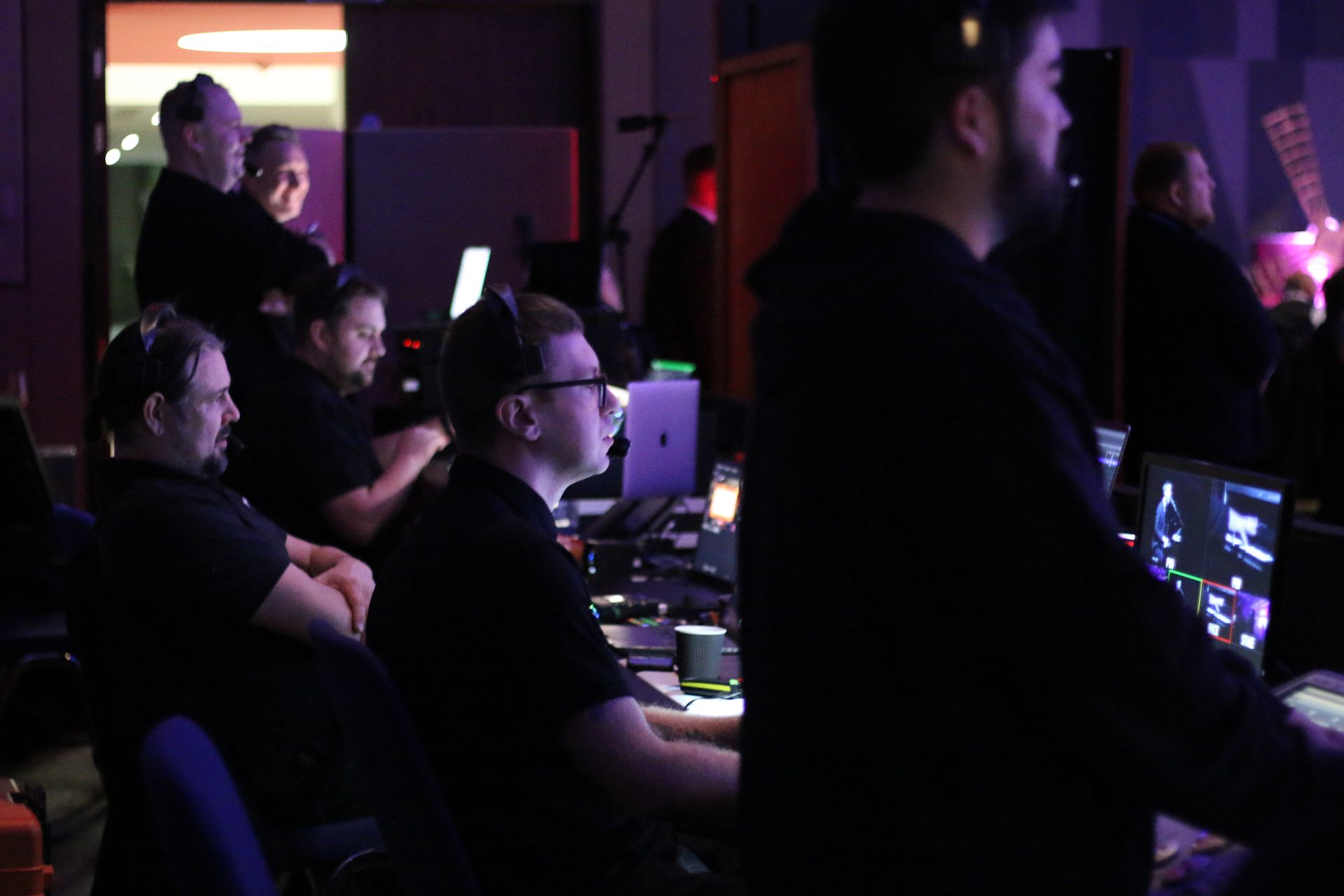 People in a darkened room with screens and equipment, possibly a control room. Most are focused on their work.