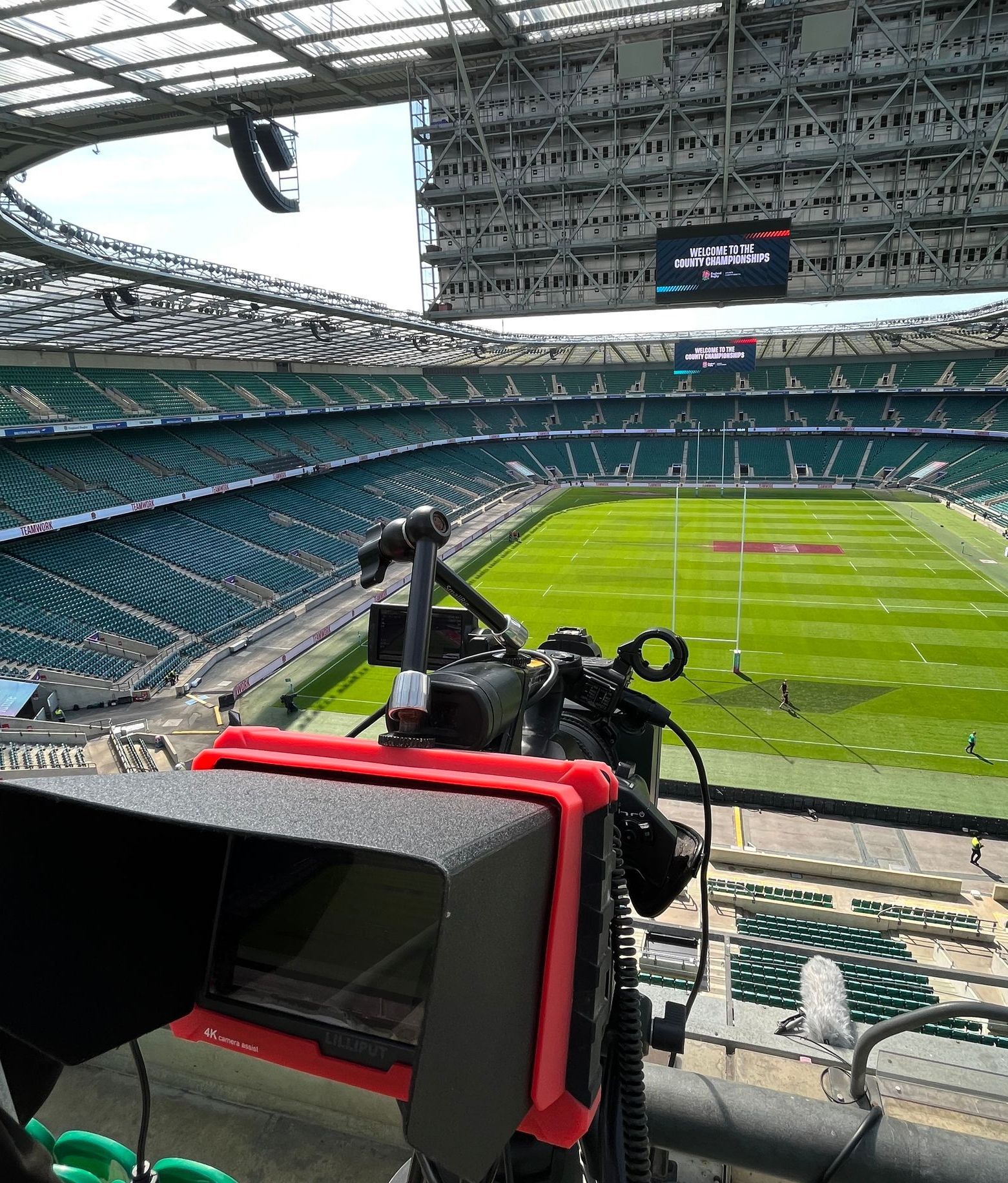 Camera set up, angled towards the green field of a rugby stadium, with seating and overhead lights visible.