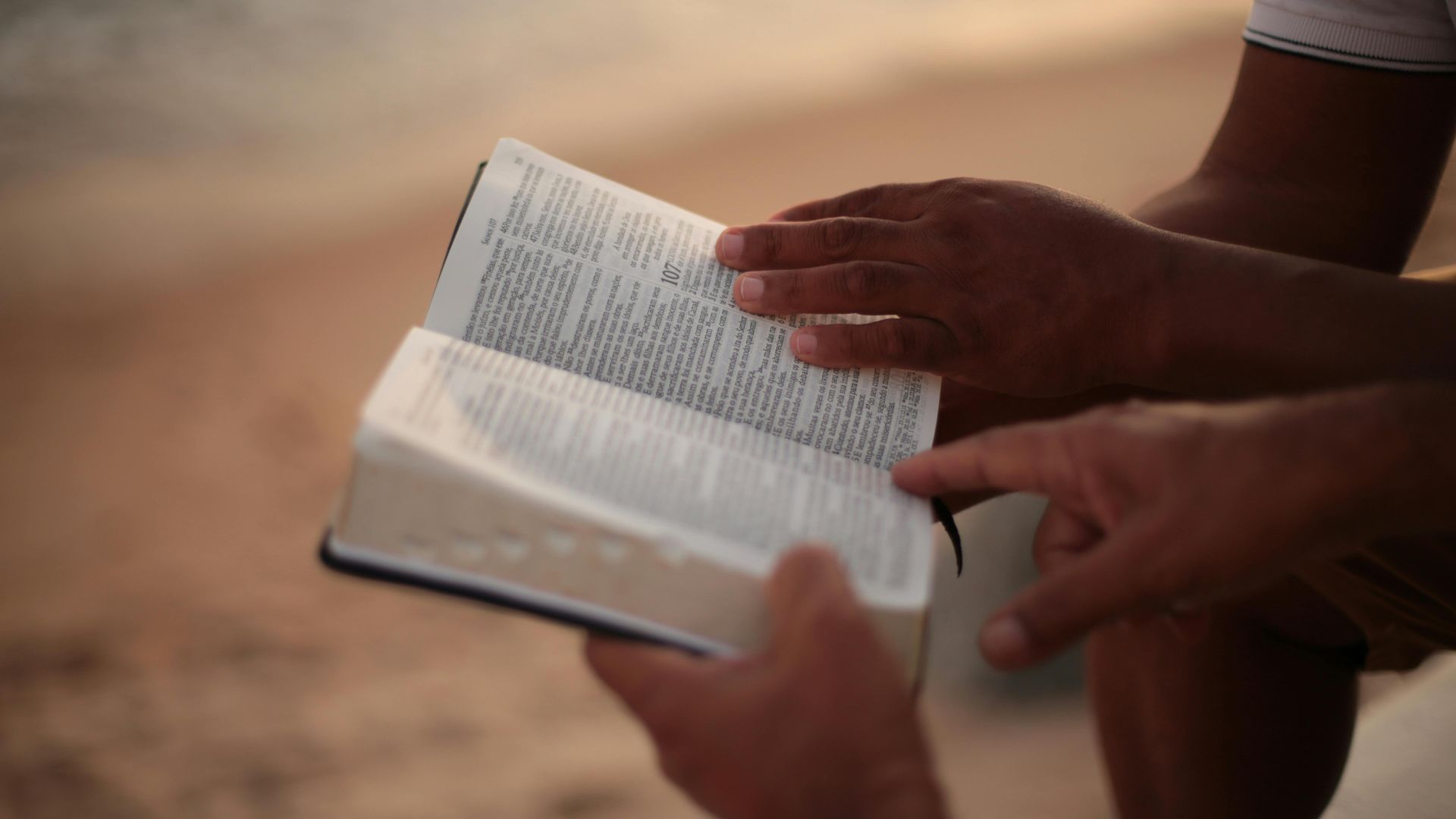 Hands holding an open book outdoors on a sandy background.