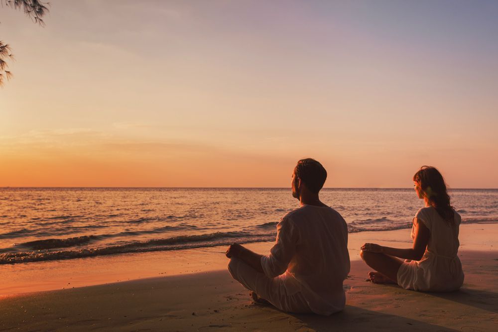 Couple Sitting In A Lotus Position On The Beach At Sunset — Kym Connolly Wellness Coach in Svensson Heights, QLD