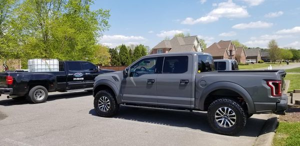 A gray Ford Raptor pickup truck parked on a driveway, with a black truck and trailer in the background.